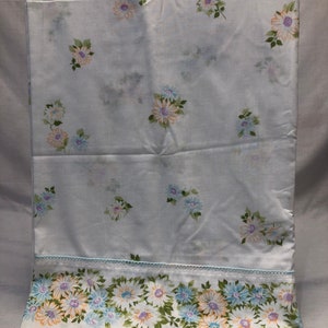 Puede incluir: Sábana bajera blanca con un patrón floral con flores azules, naranjas y moradas. La sábana tiene un borde decorativo con un patrón floral repetido.