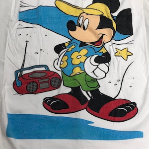 May include: A white beach towel with a cartoon image of Mickey Mouse wearing a yellow hat, blue shirt with yellow flowers, green shorts, and red sandals. Mickey is standing next to a red boombox with a yellow antenna. The background is white with blue water and a white starfish.