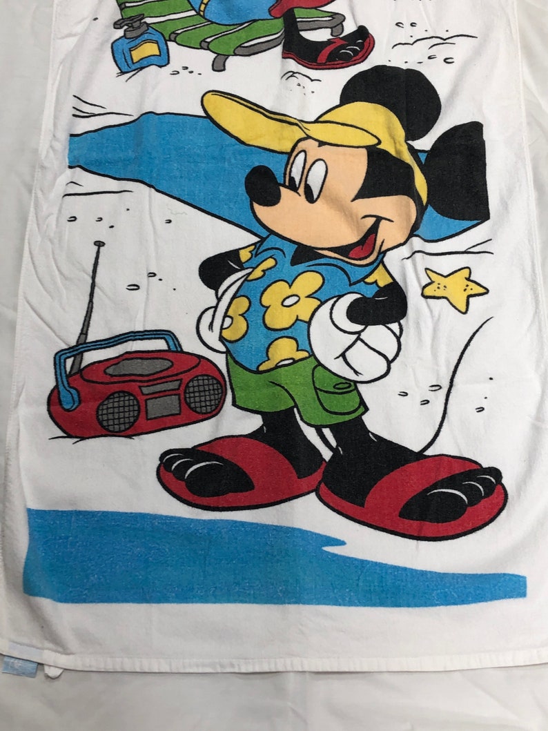 May include: A white beach towel with a cartoon image of Mickey Mouse wearing a yellow hat, blue shirt with yellow flowers, green shorts, and red sandals. Mickey is standing next to a red boombox with a yellow antenna. The background is white with blue water and a white starfish.