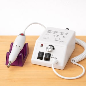 Puede incluir: Una máquina de taladro de uñas blanca con una pieza de mano blanca en un soporte púrpura. La caja de control tiene un dial, interruptores y un indicador de polaridad. El cable es blanco y en espiral. La máquina está sobre una superficie de madera clara.