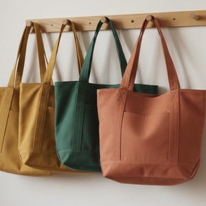 Puede incluir: Cuatro bolsas de mano de lona en amarillo mostaza, verde bosque y terracota cuelgan de un estante de madera. Cada bolsa tiene forma rectangular, un bolsillo frontal y asas largas para llevar al hombro.