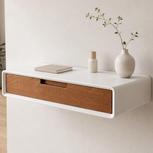 May include: A white floating wall shelf featuring a single rectangular drawer with a natural wood grain front. The shelf surface holds a small beige notebook, a cylindrical bottle with a wooden cap, and a white vase containing a green leafy branch.