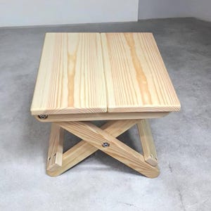 Puede incluir: Un taburete pequeño y plegable fabricado en madera de pino natural de color claro. El asiento consta de dos tablones de madera rectangulares y la base presenta un diseño de patas en cruz aseguradas con herrajes metálicos visibles.