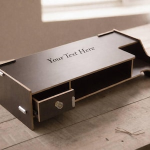 以下が含まれることがあります： 引き出しと棚が付いた、濃い茶色の木製デスクオーガナイザー。「Your Text Here」の文字が上面に印刷されています。オーガナイザーは、白い鉢植えの小さな植物が置かれた木製の机の上にあります。