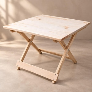 May include: A square folding table constructed from light-colored natural wood with visible grain and knots. The table features a flat top surface supported by a collapsible X-frame base with metal hardware connecting the wooden legs.