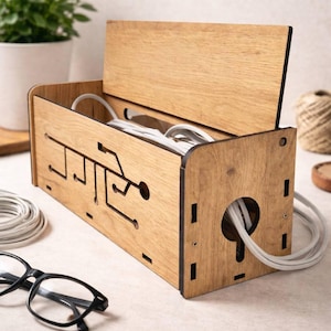 Puede incluir: Una caja organizadora de cables de madera con tapa con bisagras. La caja tiene un diseño de placa de circuito cortado en la parte delantera y un agujero grande para que pasen los cables. Cables blancos son visibles dentro. Gafas negras y una planta en el fondo.