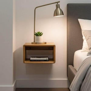 May include: A floating wooden nightstand with rounded corners mounted on a wall. A metallic desk lamp with a curved arm sits on top next to a small potted succulent. A book is placed inside the open storage compartment of the nightstand.