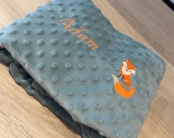 May include: A blue blanket with a dimpled texture. It has an embroidered orange fox and the name "Adam" in orange script. The blanket is folded, showing its soft, plush appearance.