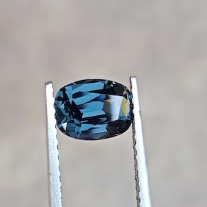May include: A faceted, oval-shaped gemstone with a deep teal color is held by silver tweezers. The gem's surface reflects light, showcasing its intricate cuts and facets. The background is a soft, neutral tone, highlighting the gem's brilliance.