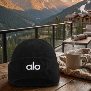 Puede incluir: Un gorro negro con el texto blanco "alo" se muestra en una mesa de madera. El fondo presenta una vista panor&aacute;mica de monta&ntilde;as y un valle. Una taza de caf&eacute; est&aacute; sobre una manta de punto.