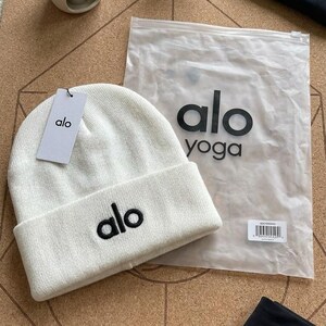 Puede incluir: Un gorro blanco con el nombre de la marca "alo" bordado en negro. El gorro tiene un borde doblado y una etiqueta con el logotipo "alo". Tambi&eacute;n se ve una bolsa de pl&aacute;stico transparente con "alo yoga" en negro, junto con leggings negros.