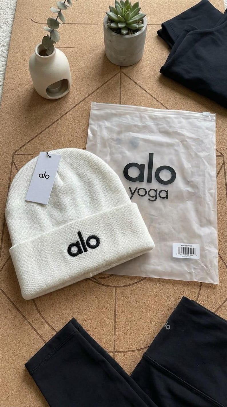 Puede incluir: Un gorro blanco con el nombre de la marca "alo" bordado en negro. El gorro tiene un borde doblado y una etiqueta con el logotipo "alo". Tambi&eacute;n se ve una bolsa de pl&aacute;stico transparente con "alo yoga" en negro, junto con leggings negros.