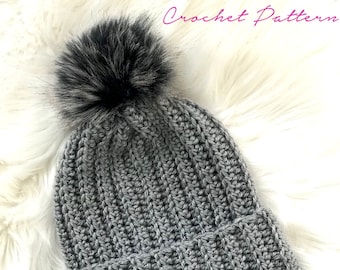 Könnte beinhalten: Eine graue, gerippte Strickmütze mit einem Kunstfell-Bommel. Die Mütze ist aus weichem, strukturiertem Garn gefertigt und hat einen umgeschlagenen Saum. Der Text "Crochet Pattern" ist oben rechts sichtbar. Die Mütze wird auf einer weißen, flauschigen Oberfläche präsentiert.