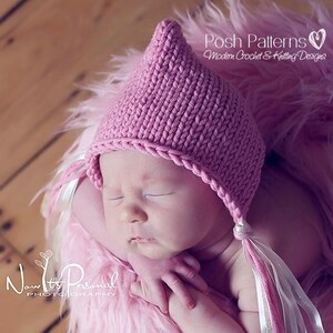 May include: A pink knitted baby hat with a pointed top and long ties. The hat is worn by a sleeping baby on a pink fluffy blanket.
