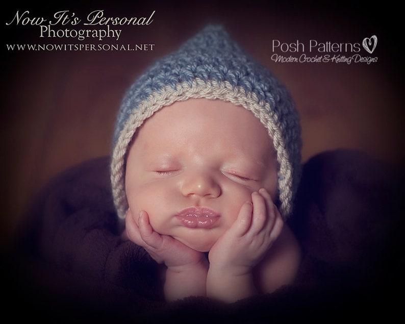 May include: A sleeping baby wearing a blue and white crocheted hat. The hat has a pointed top and a white brim. The baby is wearing a brown blanket.
