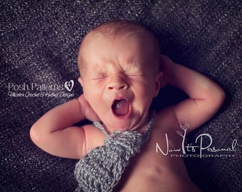 May include: A baby wearing a gray crocheted bow tie, with their mouth open in a yawn. The baby is lying on a dark gray textured surface.