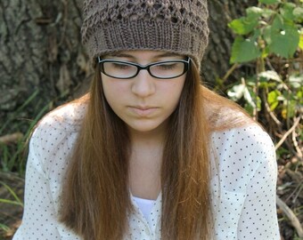 Könnte beinhalten: Eine braune Strickmütze mit einem Spitzenmuster. Die Mütze wird von einer Person mit langen braunen Haaren und Brille getragen. Die Person trägt ein weißes Hemd mit schwarzen Punkten.