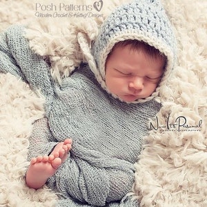 May include: A newborn baby wearing a light blue crocheted hat with a white trim. The hat has a pointed top and a button on the side. The baby is sleeping on a white fluffy blanket.
