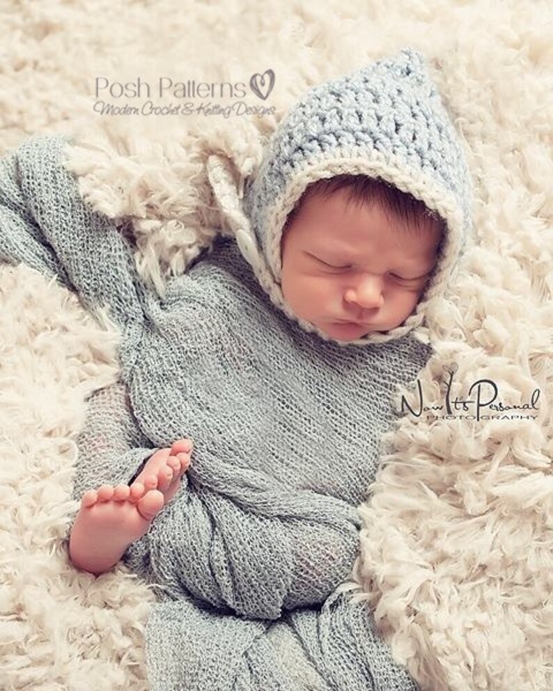 May include: A newborn baby wearing a light blue crocheted hat with a white trim. The hat has a pointed top and a button on the side. The baby is sleeping on a white fluffy blanket.
