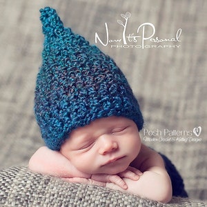 May include: A newborn baby wearing a blue crocheted hat with a pointed top. The baby is sleeping with their arms crossed in front of them. The baby is lying on a textured grey surface.