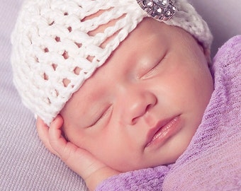 Könnte beinhalten: Ein schlafendes Baby trägt eine weiße Häkelmütze mit einem glitzernden Strassstein-Verzierungen. Das Baby liegt auf einem violetten Stoffhintergrund.