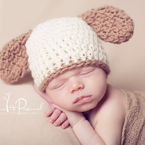 May include: A newborn baby wearing a white and brown crocheted hat with floppy ears. The baby is sleeping peacefully with their hands tucked under their chin.