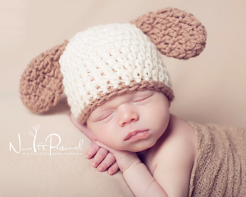 May include: A newborn baby wearing a white and brown crocheted hat with floppy ears. The baby is sleeping peacefully with their hands tucked under their chin.