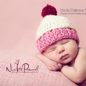 May include: A pink and white crocheted baby hat with a red pom-pom on top. The hat is on a pink blanket and a baby is sleeping on the blanket.