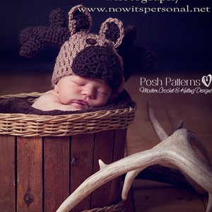 May include: A newborn baby wearing a brown and tan crocheted moose hat, sleeping in a brown wicker basket. A set of white antlers are lying on the floor next to the basket.