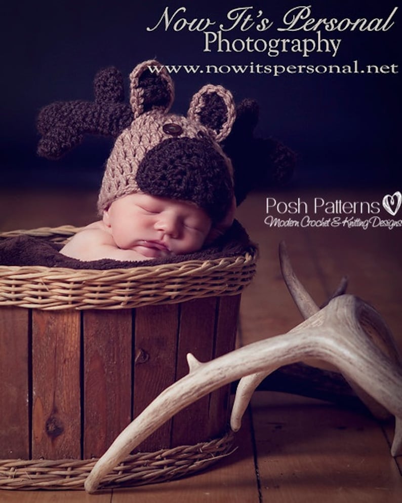 May include: A newborn baby wearing a brown and tan crocheted moose hat, sleeping in a brown wicker basket. A set of white antlers are lying on the floor next to the basket.