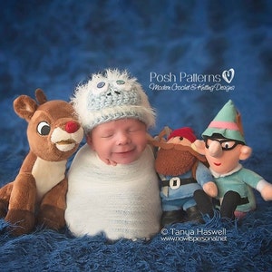 May include: A baby wearing a blue and white crocheted snowman hat is surrounded by plush toys, including a reindeer, a gnome, and an elf. The background is a blue fuzzy surface. The text "Posh Patterns Modern Crochet & Knitting Designs" is visible in the image.