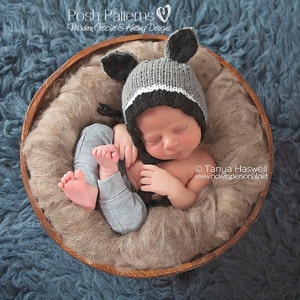 May include: A newborn baby wearing a grey and black knitted animal hat with ears, sleeping in a brown wooden bowl lined with beige faux fur.