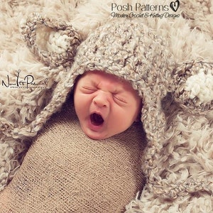 May include: A newborn baby wearing a crocheted bear hat with floppy ears. The hat is beige and white with a textured stitch. The baby is swaddled in a beige blanket and is yawning.