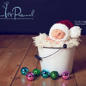 Puede incluir: Un beb&eacute; durmiendo con un gorro de Pap&aacute; Noel de crochet rojo y blanco con un pomp&oacute;n blanco en la parte superior, acostado en un cubo blanco con una manta blanca peluda. El cubo est&aacute; en un suelo de madera con cascabeles coloridos esparcidos a su alrededor.