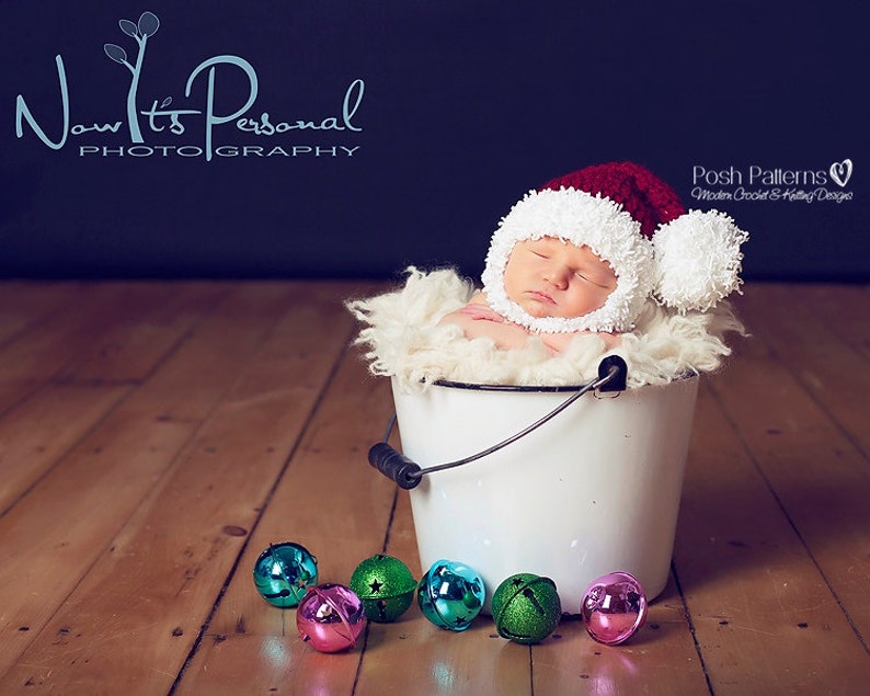 Puede incluir: Un beb&eacute; durmiendo con un gorro de Pap&aacute; Noel de crochet rojo y blanco con un pomp&oacute;n blanco en la parte superior, acostado en un cubo blanco con una manta blanca peluda. El cubo est&aacute; en un suelo de madera con cascabeles coloridos esparcidos a su alrededor.