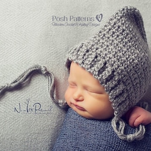 Op de afbeelding: Een grijze gehaakte baby muts met een lange, gebonden kwast. De muts heeft een puntige bovenkant en bedekt de oren. De muts wordt gedragen door een slapende baby.