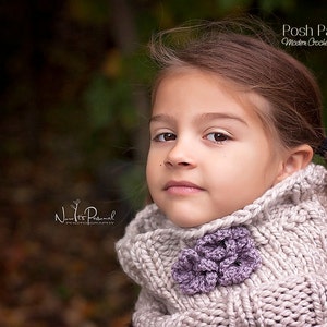 May include: A young girl wearing a light beige knitted cowl neck scarf with a purple flower embellishment. The scarf is made with a chunky yarn and has a textured stitch pattern.