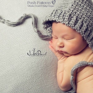 May include: A gray crocheted baby bonnet with a long tie. The bonnet is worn by a sleeping baby on a gray textured background.