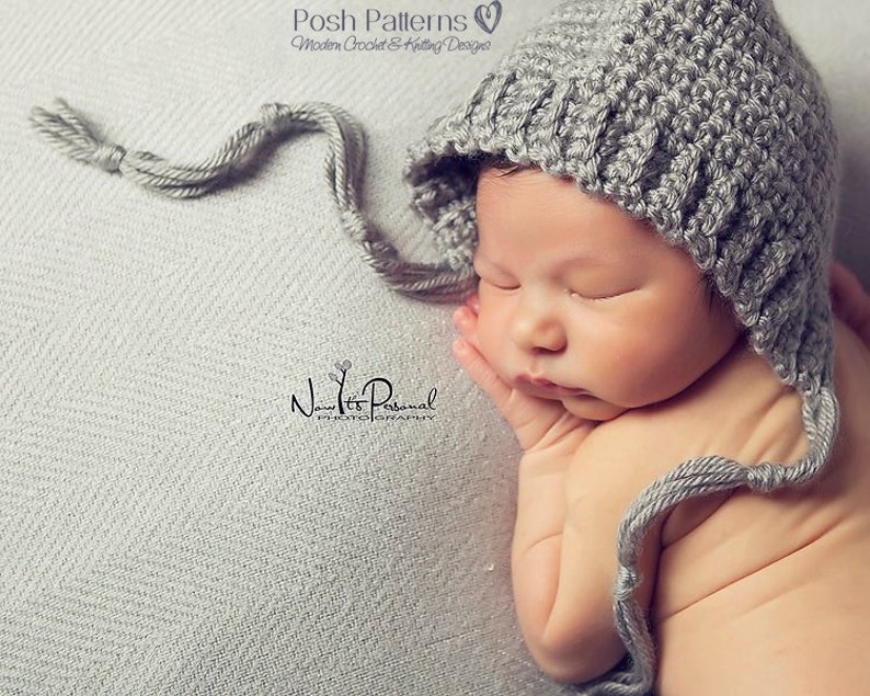 May include: A gray crocheted baby bonnet with a long tie. The bonnet is worn by a sleeping baby on a gray textured background.