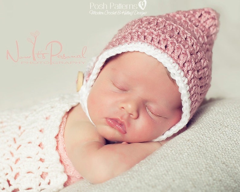 May include: A sleeping baby wearing a pink and white crocheted hat with a button on the side. The baby is lying on a white blanket.