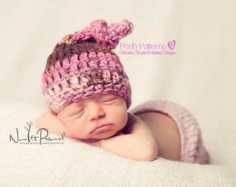 May include: A newborn baby wearing a pink and brown crocheted hat with a knotted top. The hat is made with a textured stitch and has a soft, cozy look. The baby is sleeping peacefully on a white blanket.