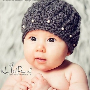 May include: A gray crocheted baby hat with a textured pattern and white pearl embellishments. The hat is worn by a baby with a light complexion and large eyes.