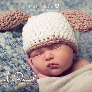 May include: A newborn baby wearing a white and brown crocheted hat with floppy ears. The hat has a brown brim and brown ears. The baby is sleeping on a blue and white patterned blanket.