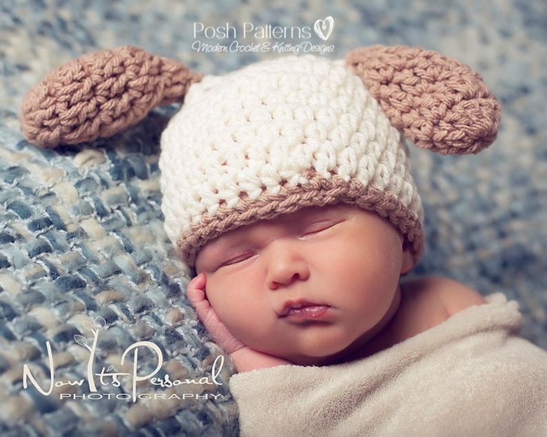 May include: A newborn baby wearing a white and brown crocheted hat with floppy ears. The hat has a brown brim and brown ears. The baby is sleeping on a blue and white patterned blanket.