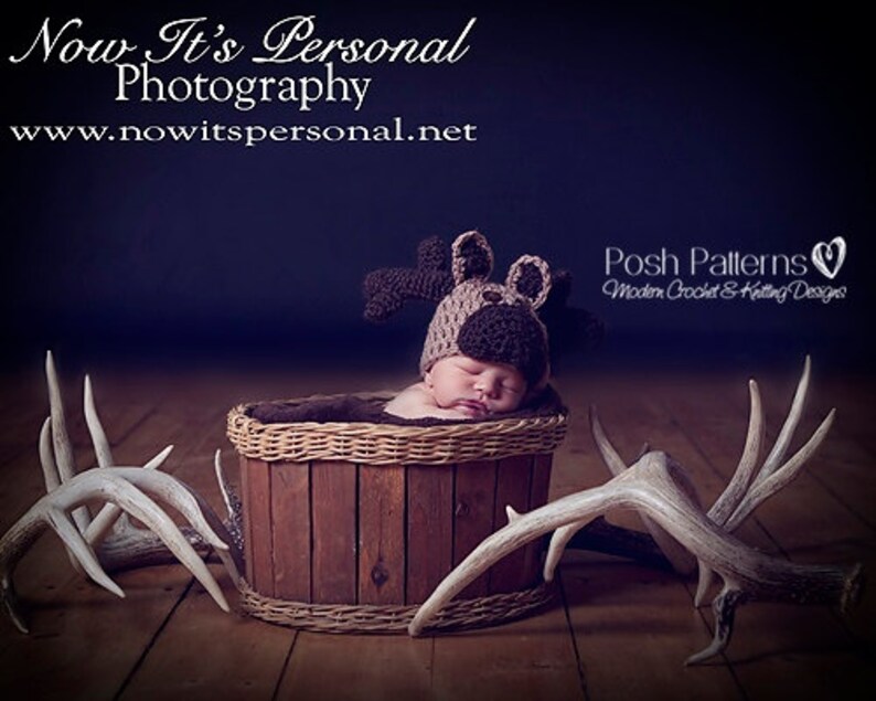 May include: A newborn baby wearing a brown crocheted moose hat is sleeping in a wooden basket. Two white antlers are on either side of the basket. The image is captioned "Now It's Personal Photography www.nowitspersonal.net" and "Posh Patterns Modern Crochet & Knitting Designs".