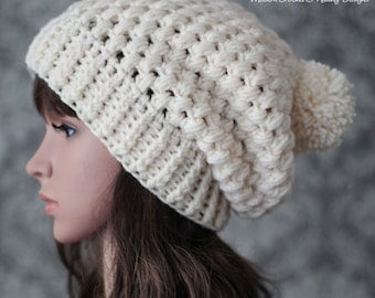 Puede incluir: Un gorro de punto blanco con un pompón en la parte superior. El gorro es holgado y tiene un patrón de punto texturizado. El gorro lo lleva puesto una persona con el pelo largo y castaño.