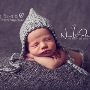May include: A gray knitted baby bonnet with a pointed top and a long tassel. The bonnet is worn by a sleeping baby on a gray textured blanket.