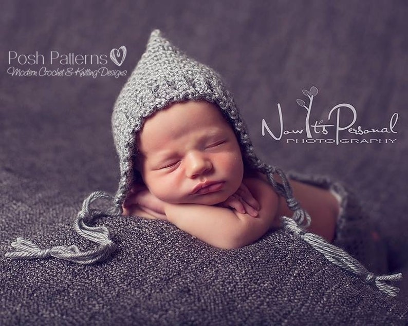 May include: A gray knitted baby bonnet with a pointed top and a long tassel. The bonnet is worn by a sleeping baby on a gray textured blanket.