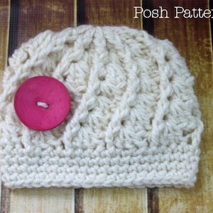 Könnte beinhalten: Eine weiße Häkelmütze mit einem rosa Knopf. Die Mütze hat ein strukturiertes, gekreuztes Strickmuster.