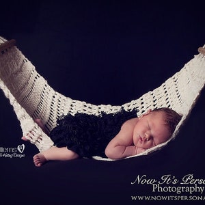 May include: A newborn baby sleeping in a white crocheted hammock. The baby is wearing a black tutu and is lying on its side with its arms tucked in. The hammock is hanging from a dark background.